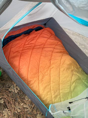 Sleeping bag inside a tent, showing a gradient pattern, set on a forest floor with pine needles