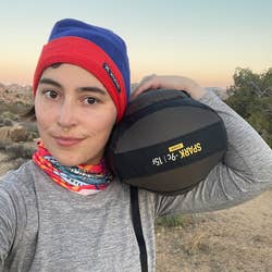 Person outdoors holding a sleeping bag, wearing a beanie and neck gaiter, posing for a selfie in a desert landscape