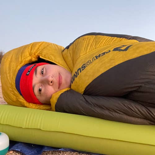 Person lying on a sleeping mat outdoors, wrapped in a yellow and brown sleeping bag, wearing a red beanie