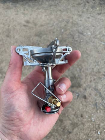 Hand holding a compact camping stove with a metal burner, piezo igniter button, and control valve
