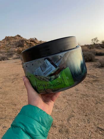 Person holding a transparent container filled with snacks and a lid, outdoors in a desert setting. Suitable for travel or camping