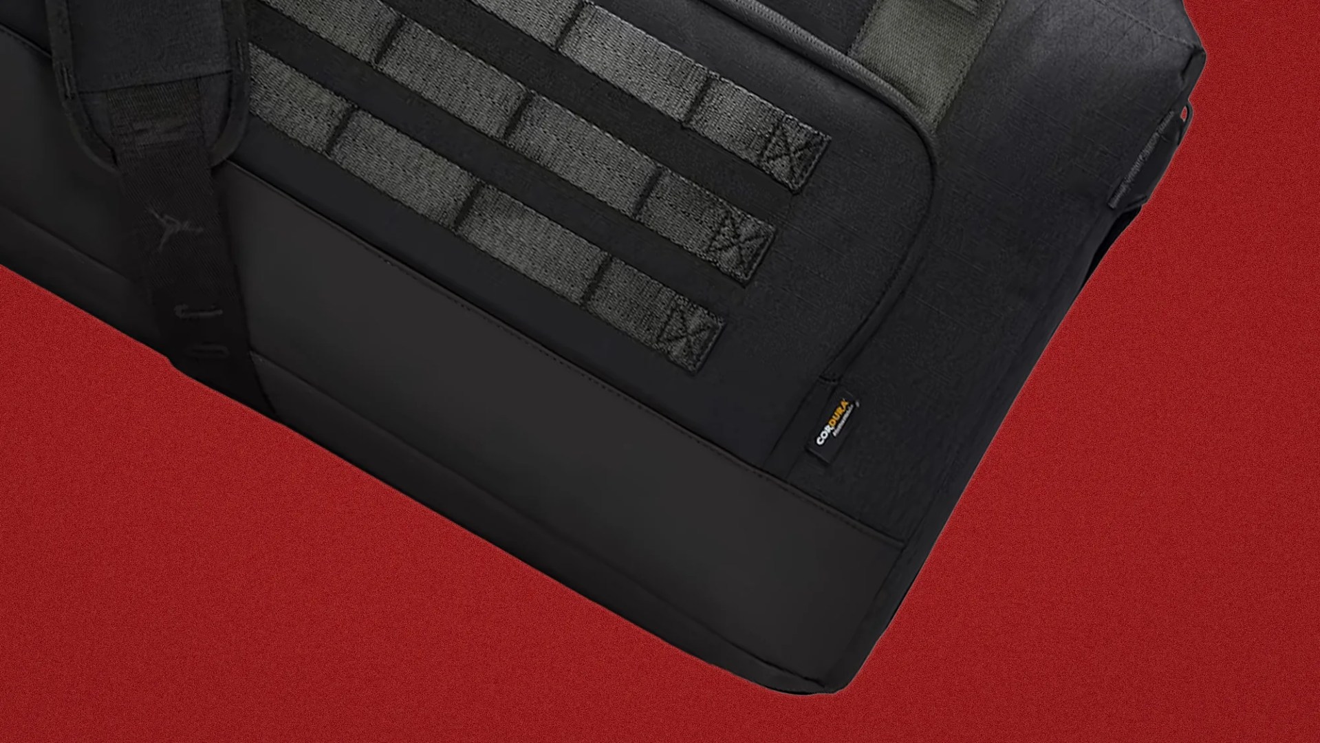 Close-up of a black tactical bag with MOLLE webbing and a small Cordura fabric label on a red background.