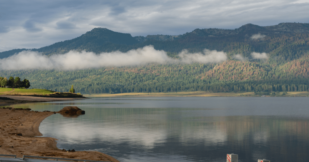 Scenic view of Idaho's Lake Cascade surrounded by mountains and trees, with a sandy shore in the foreground and mist in the distance under a cloudy sky.