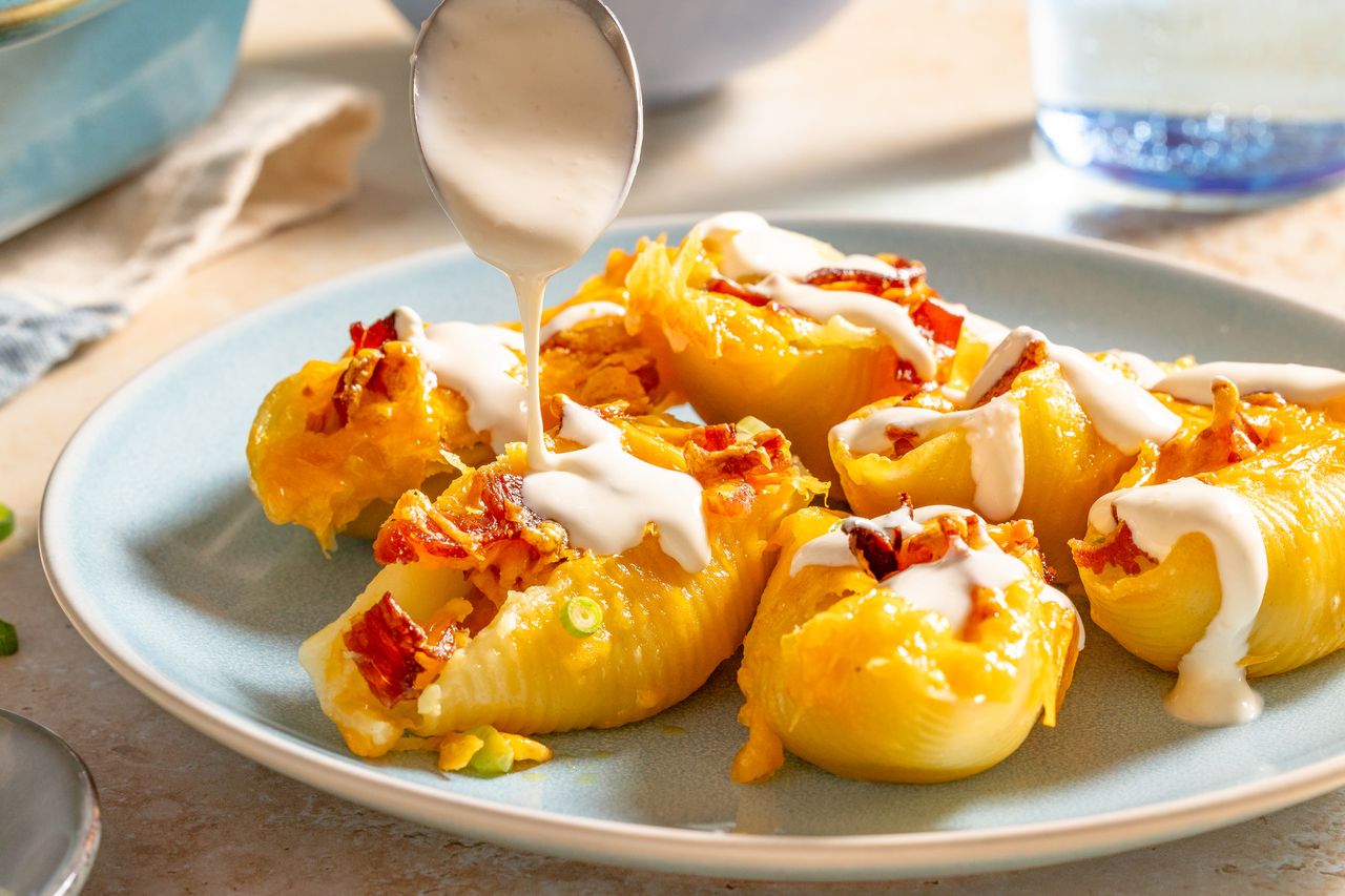 Stuffed pasta shells on a plate being drizzled with sauce topped with cheese and crumbled bacon