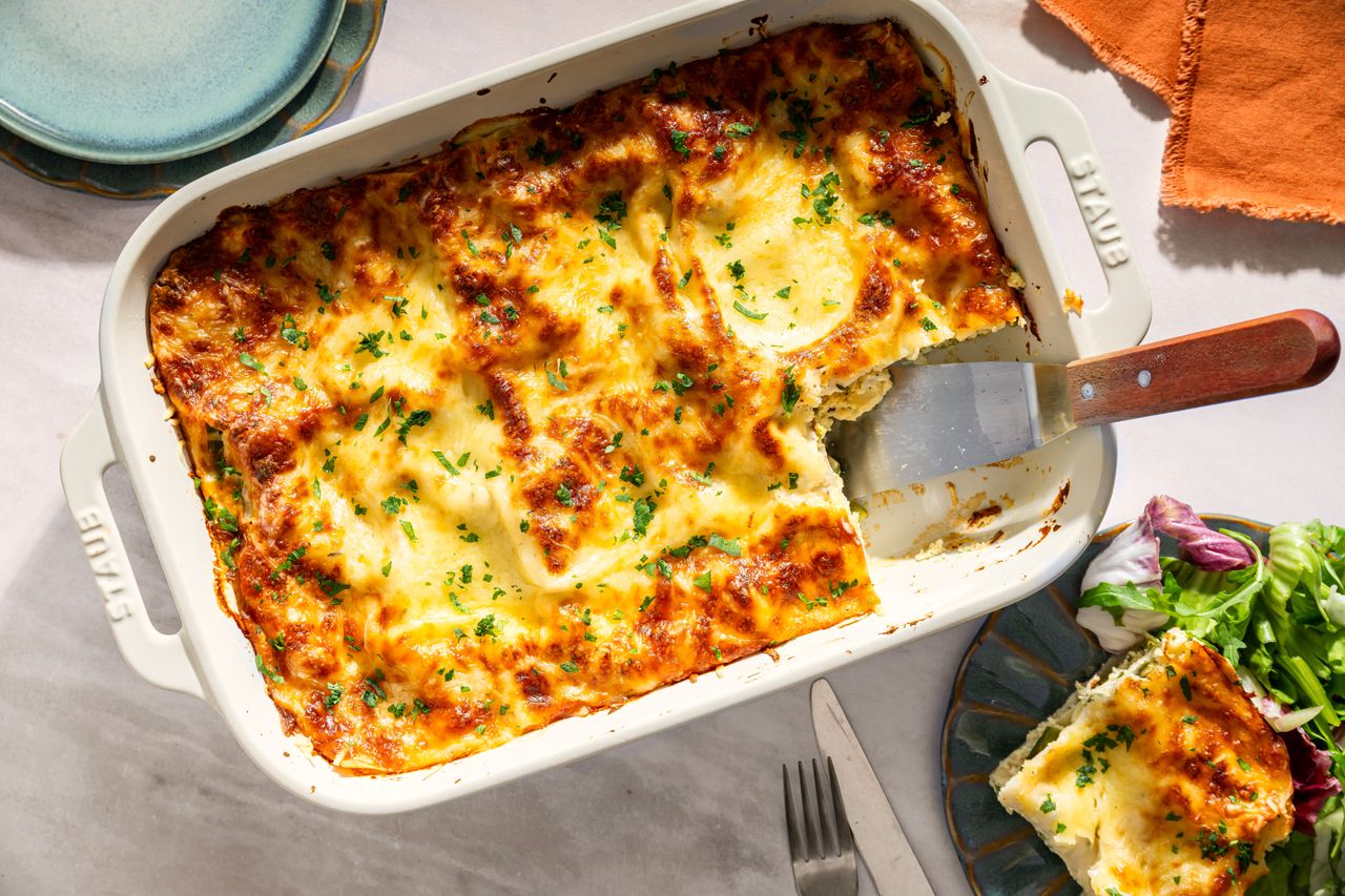 A baked lasagna in a casserole dish with a serving removed served alongside a fork and a portion of salad
