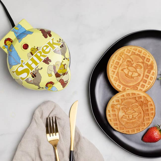 Shrek-themed waffle maker with Shrek and Donkey images, two waffles featuring Shrek's face on a black plate, with a strawberry garnish