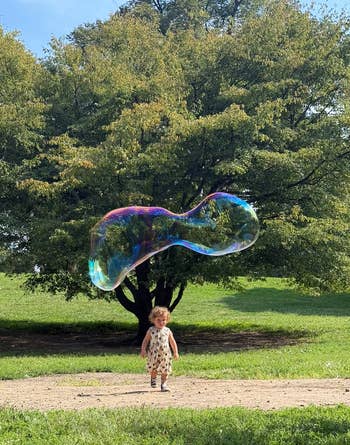 Child joyfully chasing a large bubble in a park, with green trees in the background