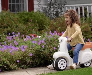 A young child with curly hair rides a white scooter-like toy on a sidewalk in front of a house with a flower garden