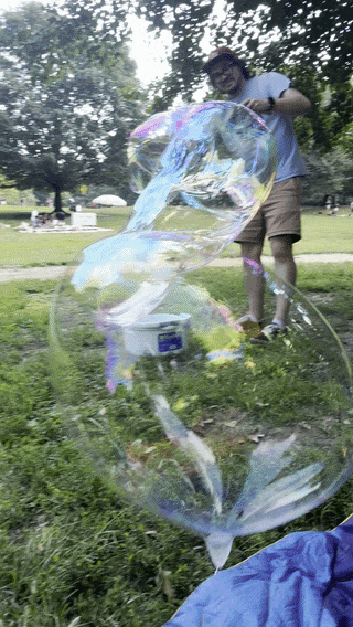 Person making large soap bubbles in a park setting, with trees and picnic area in the background