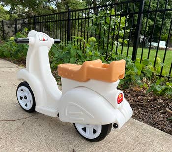 White and orange toy scooter with three wheels, placed on a sidewalk next to a garden with lush greenery