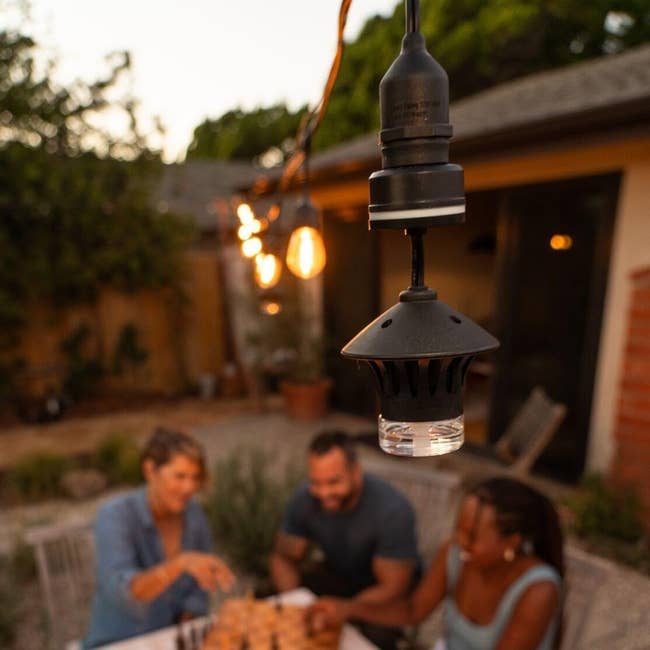 Three people enjoy a meal at an outdoor table with hanging pendant lights, creating a cozy ambiance suitable for entertaining