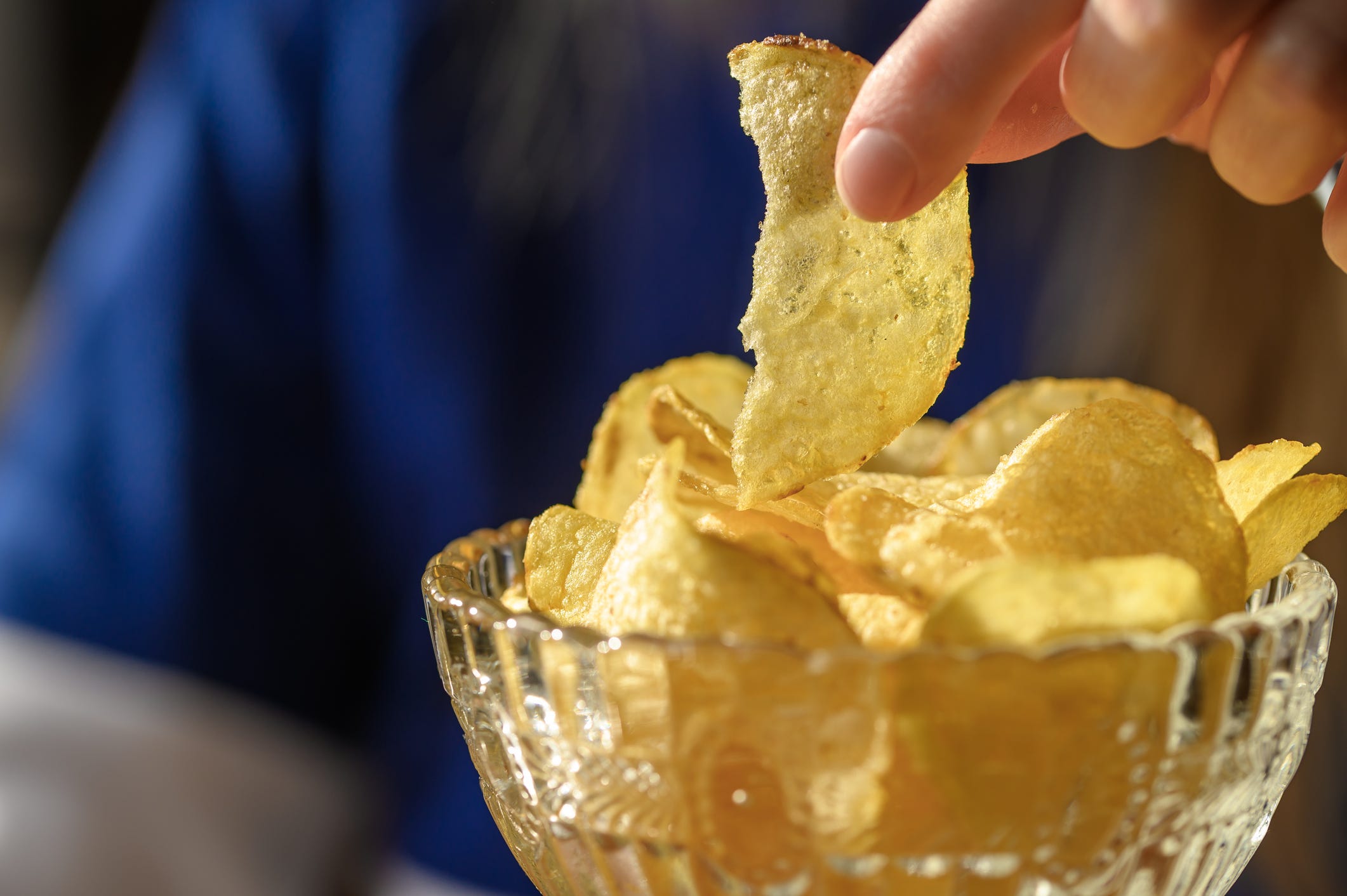 A hand grabs a thin potato chip from a glass bowl.