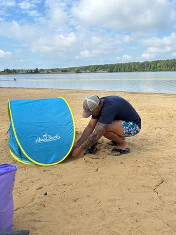 Person sets up a pop-up beach tent on sandy shore by a lake, wearing a cap, dark shirt, and floral shorts