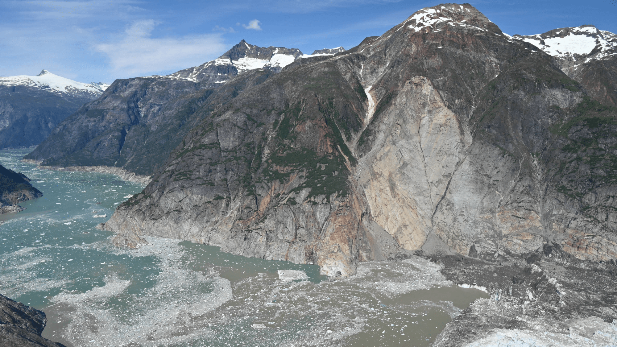 An aerial photo of the August 10, 2025 landslide taken from across the fjord during a U.S. Geological Survey field reconnaissance overflight on August 13, 2025. Note the trimline along the far side of the fjord, caused by the tsunami stripping the walls of vegetation. View direction is approximately north.