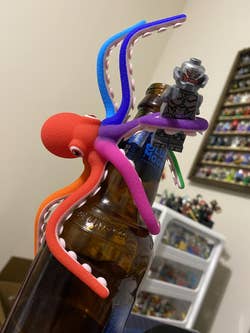 Toy octopus bottle opener on beer bottle next to toy figure. Background shows a shelf with collectible figurines
