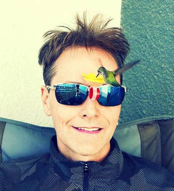 A person wearing sunglasses with a small bird drinking from a feeder attached to the glasses