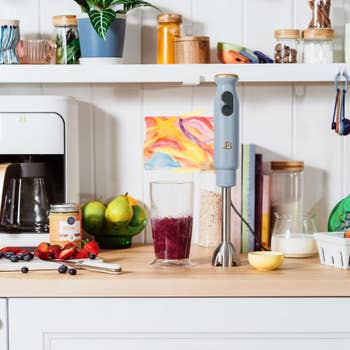 Kitchen scene with a hand blender, smoothie, fruits, and appliances on a counter. Perfect setup for making fresh, healthy drinks at home