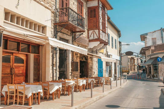 An image of a street lined with traditional buildings, some of which feature balconies and awnings. The street is empty, showcasing rows of outdoor dining tables and chairs set up in front of the establishments. The pavement is smooth, with a series of metal poles, possibly for bollards or barriers. The sky is clear, indicating a sunny day.