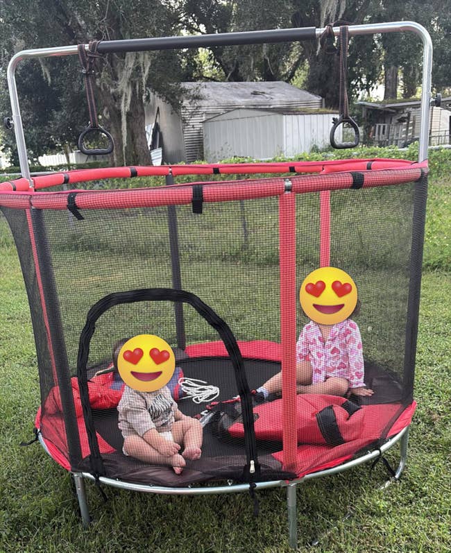 Two young children sitting inside a small outdoor trampoline with toys nearby. They appear to be enjoying their time together