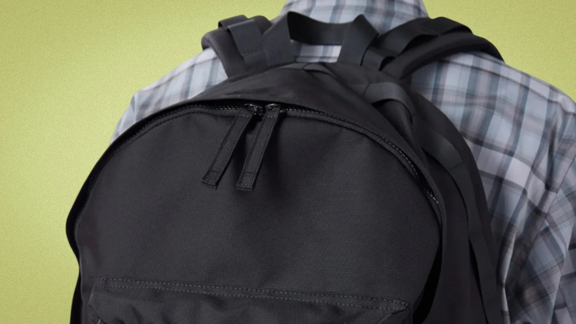 Black fabric backpack with double zipper pulls worn over a plaid shirt.