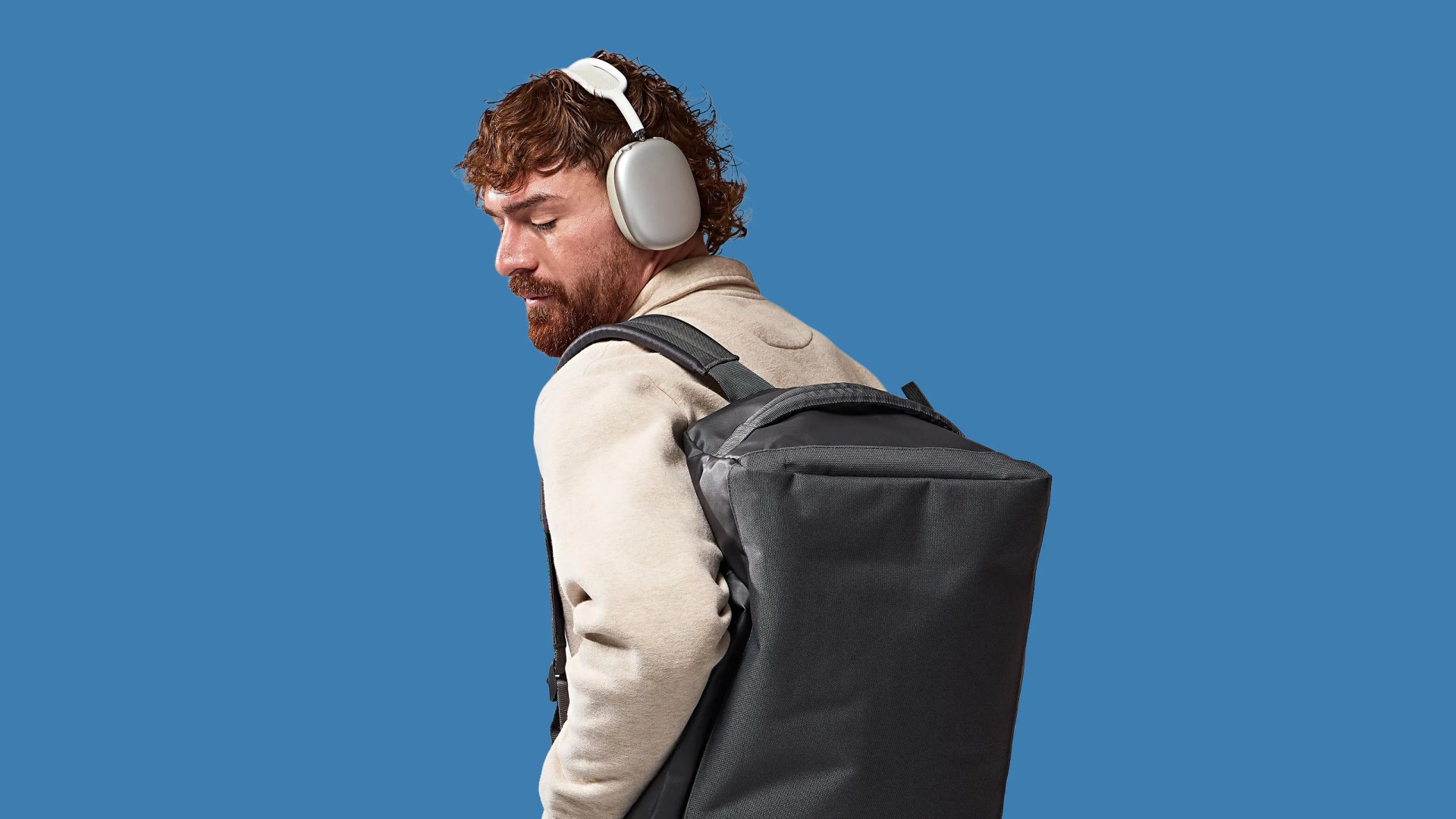 Person wearing beige jacket, silver over-ear headphones, and carrying a large black backpack against a blue background.