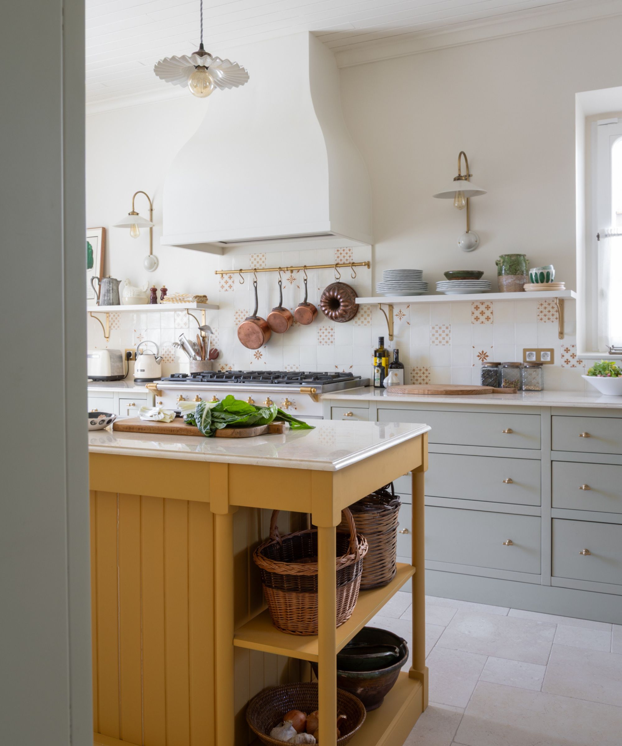 A country kitchen with pale blue cabinets, a yellow island, and a tile backsplash with open shelving and a brass pot rail