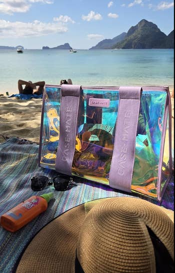 Beach scene with a transparent Case-Mate tote, sunscreen, sunglasses, and a straw hat on a colorful towel. Scenic view of sea and mountains in background