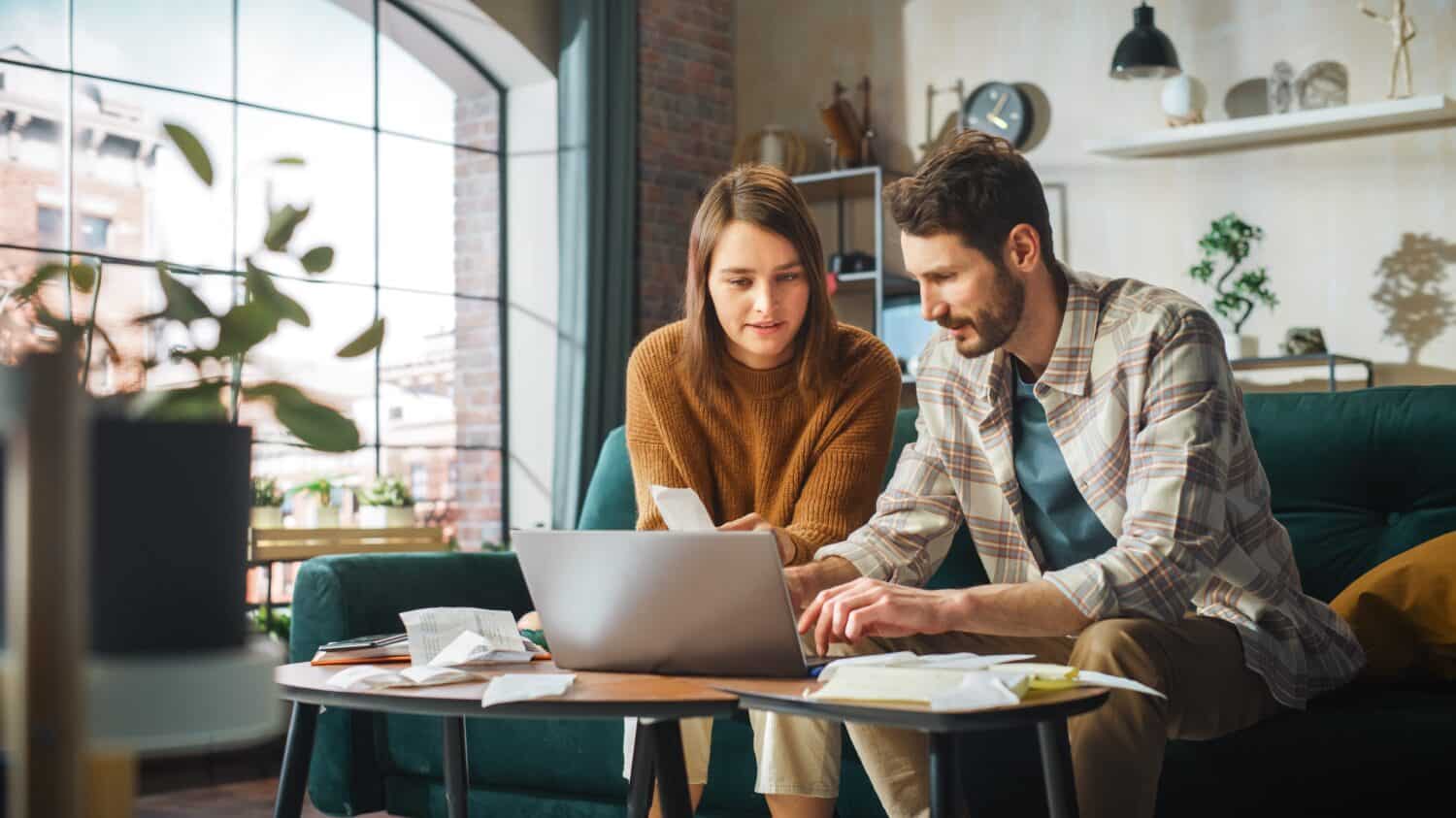A young Caucasian couple sits on a dark green sofa, focused on a silver laptop placed on a wooden coffee table. The woman, wearing a brown sweater, holds a document while the man, in a plaid shirt, points to the screen. Scattered papers and folders are on the table. The background features a bright window, plants, and a partial view of a brick wall.