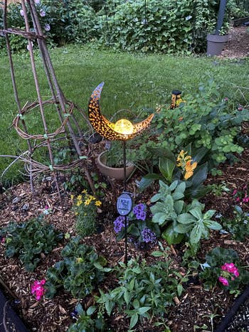 A decorative solar garden light shaped like a crescent moon stands amid vibrant flowers and greenery in a landscaped garden