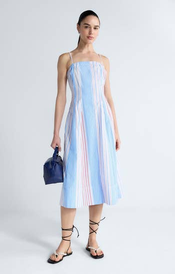 Person in a striped, spaghetti-strap midi dress, holding a small bag, wearing lace-up sandals, standing against a plain background