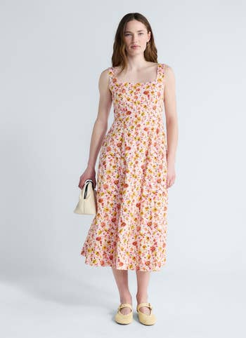 Person in a floral-patterned sleeveless dress holds a small bag, wearing casual shoes, posing for a fashion shoot