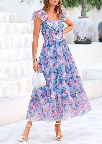 Model wearing a floral-patterned, sleeveless, tiered maxi dress with puff shoulders, holding a small white handbag