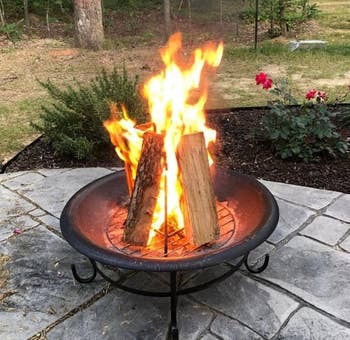 A fire pit with burning logs on a stone patio, surrounded by plants and outdoor scenery