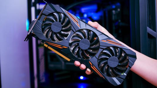 Person holding a large triple-fan graphics card in front of an open desktop gaming PC with blue internal lighting.