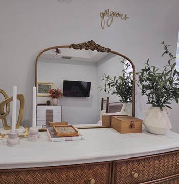 Dresser with decorative items, mirror reflecting TV and bed, potted plants, and a wall sign saying