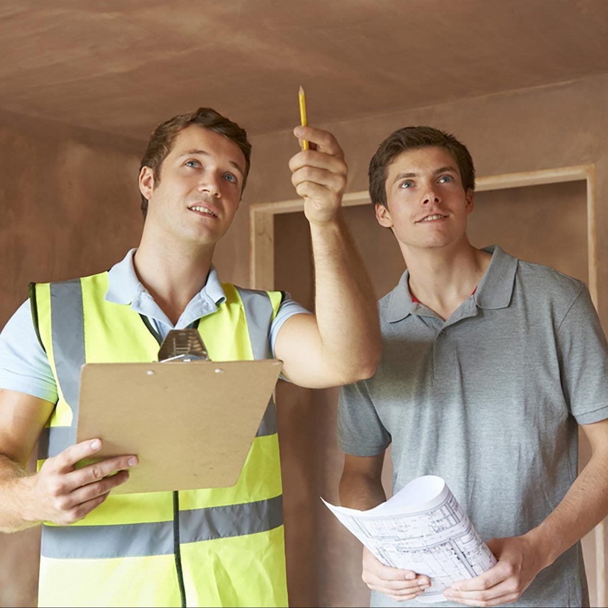 Two men discuss a construction project; one points upward holding a pencil, while the other holds blueprints in a partially finished room.