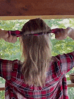 Person tying headband over long hair in outdoor setting, wearing a plaid shirt
