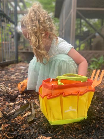 Child gardening with orange tools in a backyard, wearing a tulle dress