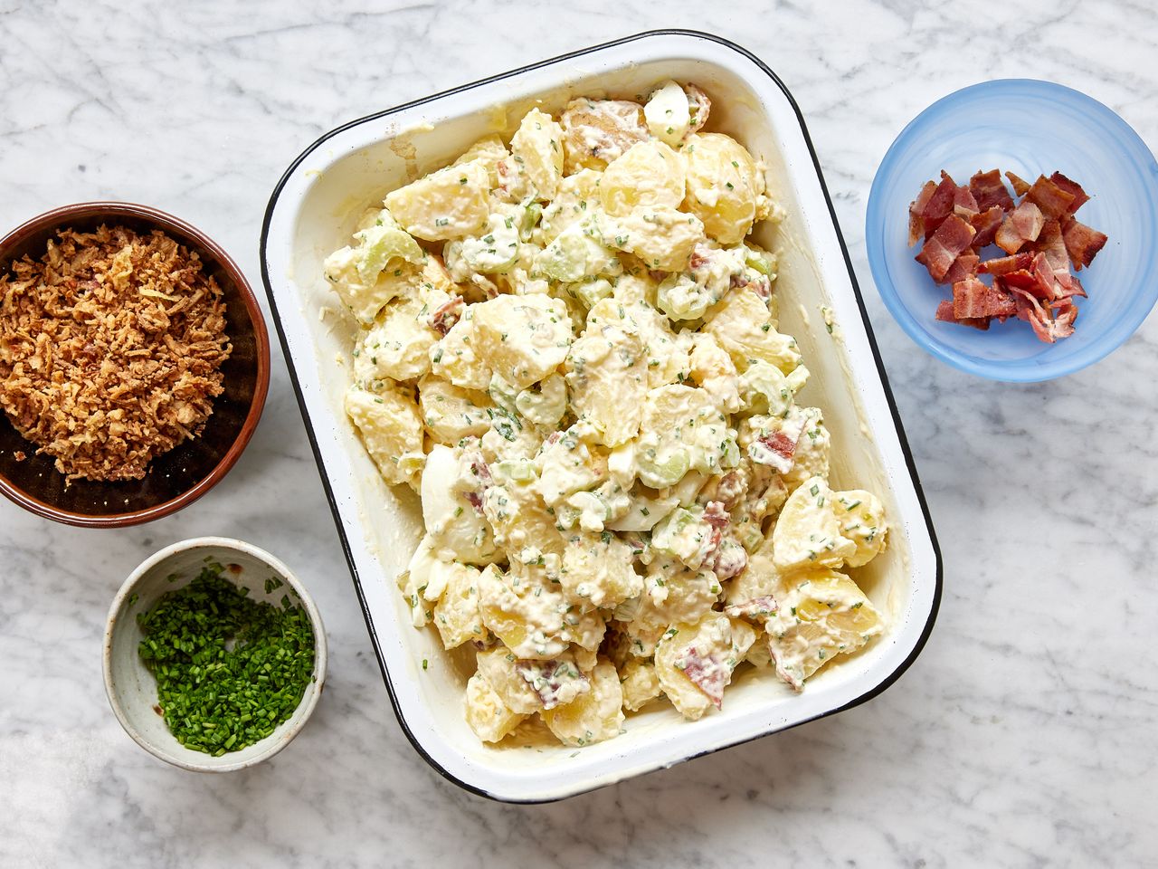 A dish of million dollar potato salad served with chopped chives, bacon, and crispy fried onions on the side