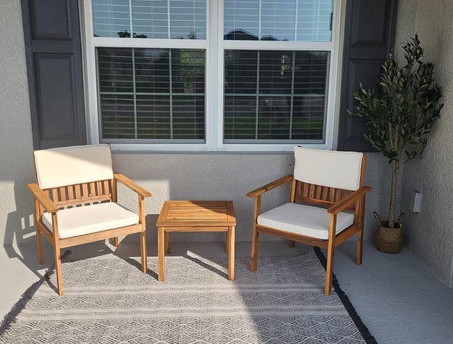 Two wooden chairs with white cushions flank a small wooden table on a porch with a potted plant and window with blinds in the background