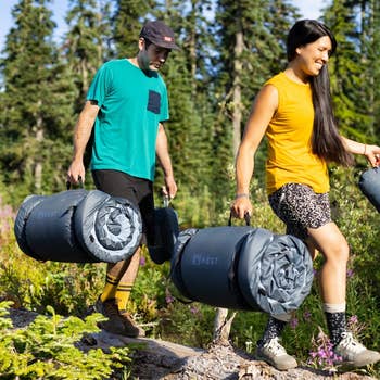 Two people hiking in the forest carry rolled-up camping gear. They wear casual outdoor clothing, and the woman has leopard-patterned shorts
