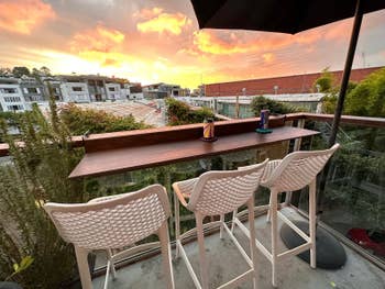 Rooftop patio with three white bar stools at a high table, overlooking a vibrant sunset sky and urban landscape