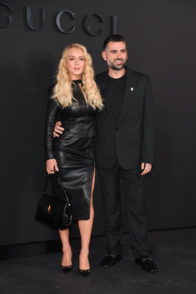 Lindsay Lohan poses for a photo with her husband Bader Shammas in front of a black Gucci background. Lohan is wearing a two-piece leather dress with a thigh-high slit and matching pointed-toe pumps, while Shammas is wearing an all-black suit over a black shirt.