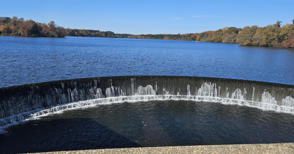 A small man-made waterfall in Parvin Lake in New Jersey, with fall foliage trees in the background.