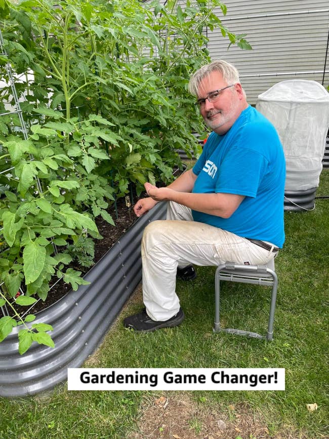 Person in a blue shirt and white pants sitting on a stool, gardening next to a raised metal planter with lush green plants