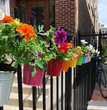 Colorful flowers in pots hang on a black fence along a sidewalk, enhancing outdoor decor with vibrant blooms