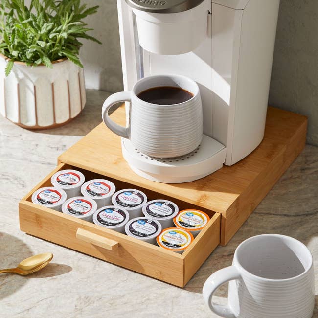 Single-serve coffee maker on a counter with a mug filled with coffee and a wooden drawer containing assorted coffee pods underneath