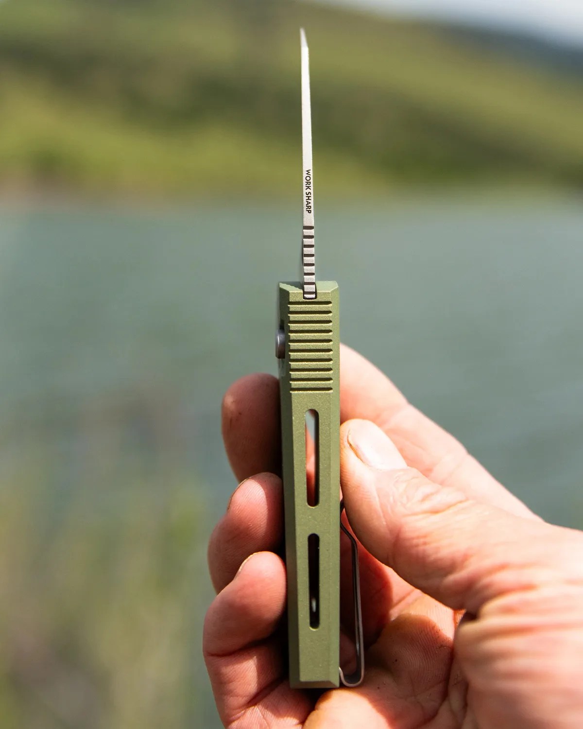 Hand holding a green folding knife with a partially serrated blade and "WORK SHARP" text on the blade.