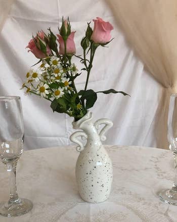 Porcelain vase with floral design holds pink roses and daisies on an elegant lace-draped table, flanked by two empty wine glasses