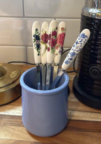 Five floral-patterned butter knives with ceramic handles stand in a blue holder on a kitchen counter